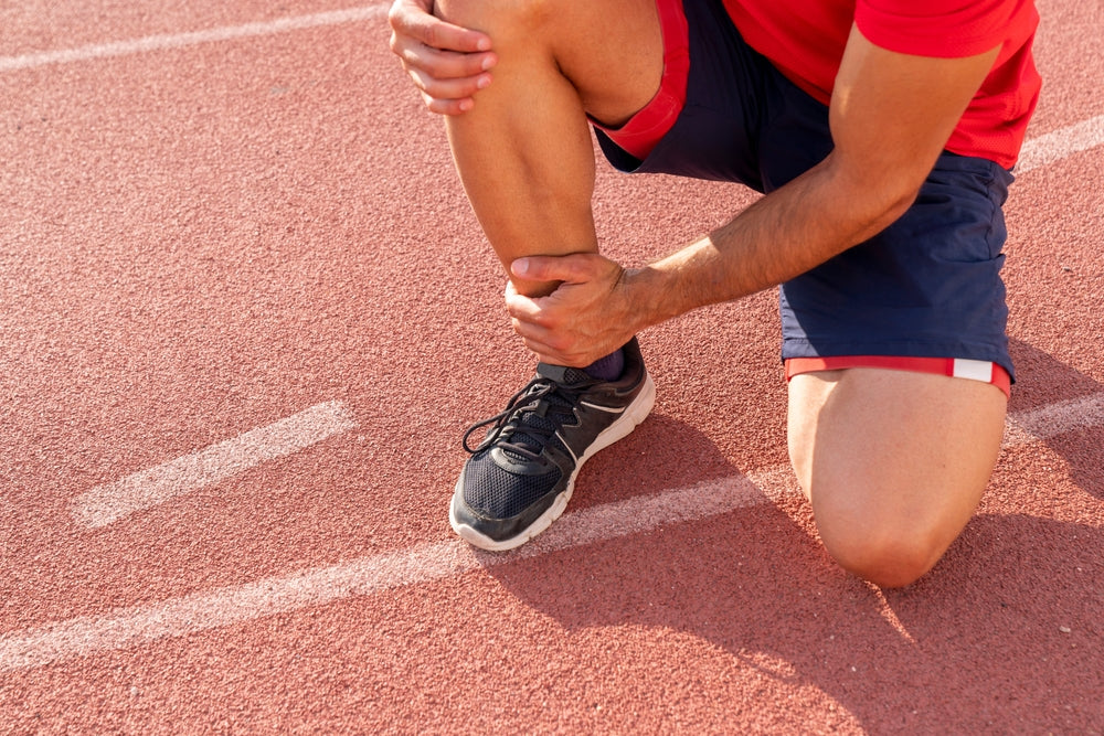 A close-up of a runner kneeling on a red running track, holding and examining their shin while wearing a red and navy athletic outfit and a black running shoe.