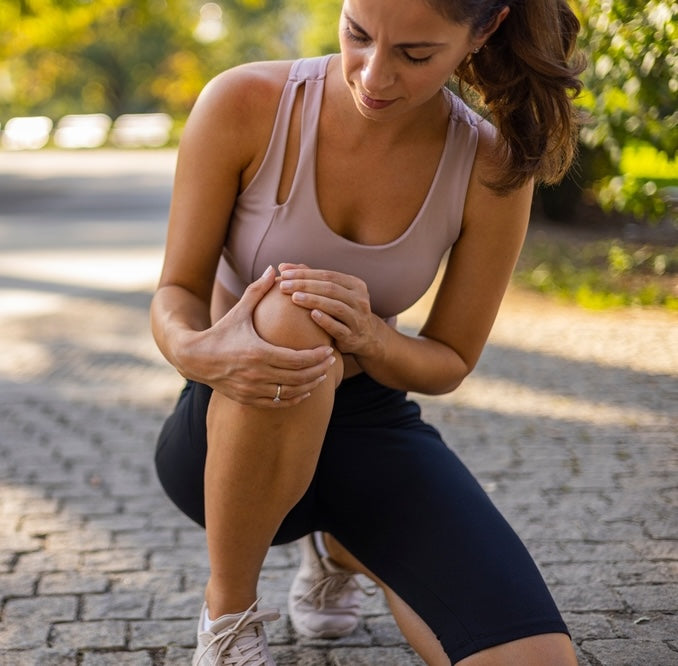 Woman in athletic wear holding her knee outdoors on a paved path.