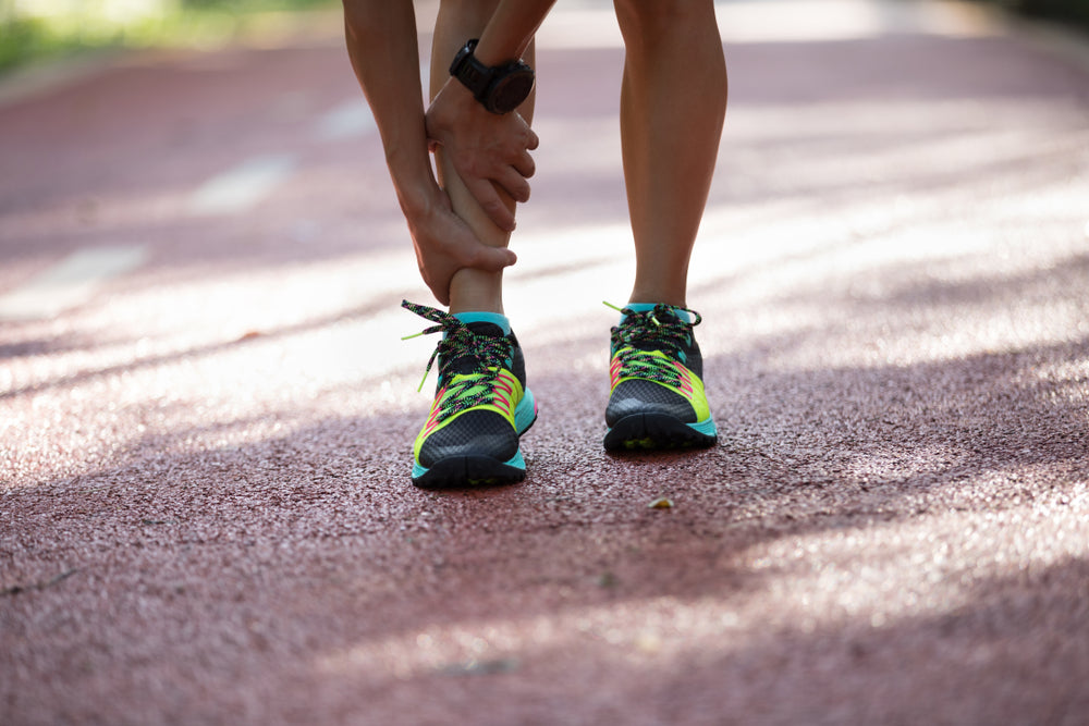 A close-up of a runner's legs and feet on a reddish-brown running track, holding their shin. They are wearing black athletic shoes with bright neon green and turquoise accents, along with a black smartwatch.