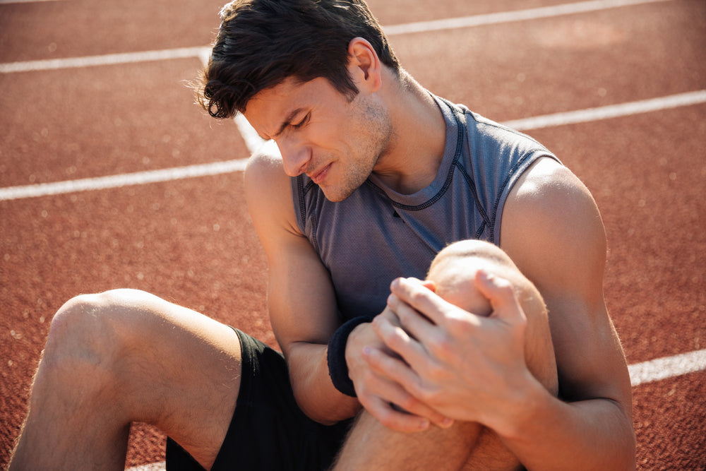 A man sitting on a red running track, holding his knee in pain wearing a gray sleeveless athletic shirt and black shorts.