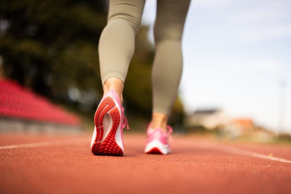 A close-up rear view of a person running on an outdoor track, wearing light olive leggings and pink running shoes with red outsole details.