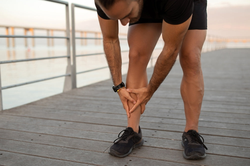 A man in black athletic wear bends over to examine his lower leg on a wooden boardwalk by the water.