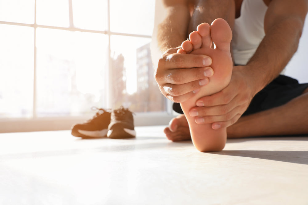 A close-up lifestyle shot of a person holding their foot, with a pair of brown athletic sneakers blurred in the background near a bright window.