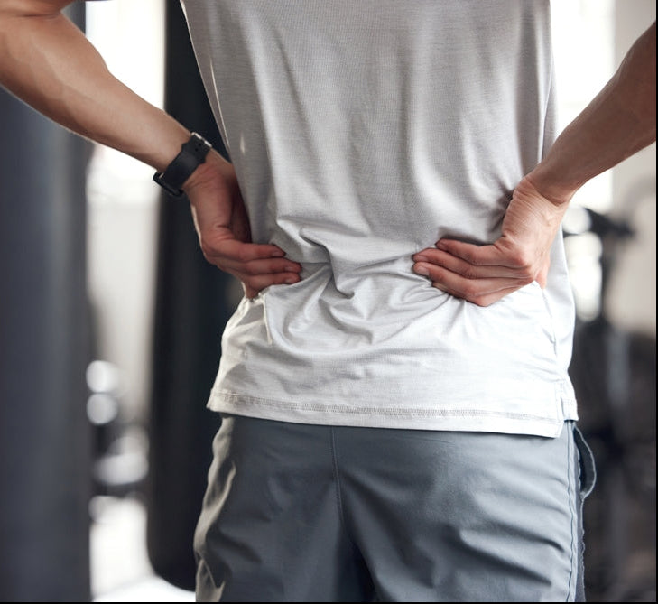A man from behind wearing a light gray t-shirt and gray athletic shorts, holding his lower back in pain.