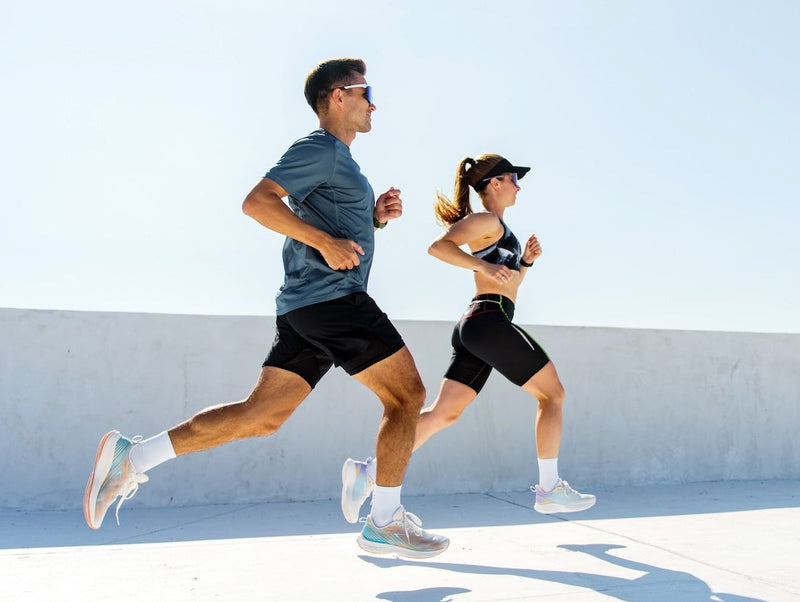 A man and a woman running side by side outdoors on a sunny day. The man wears a slate blue t-shirt, black running shorts, white socks, and light-colored running shoes.